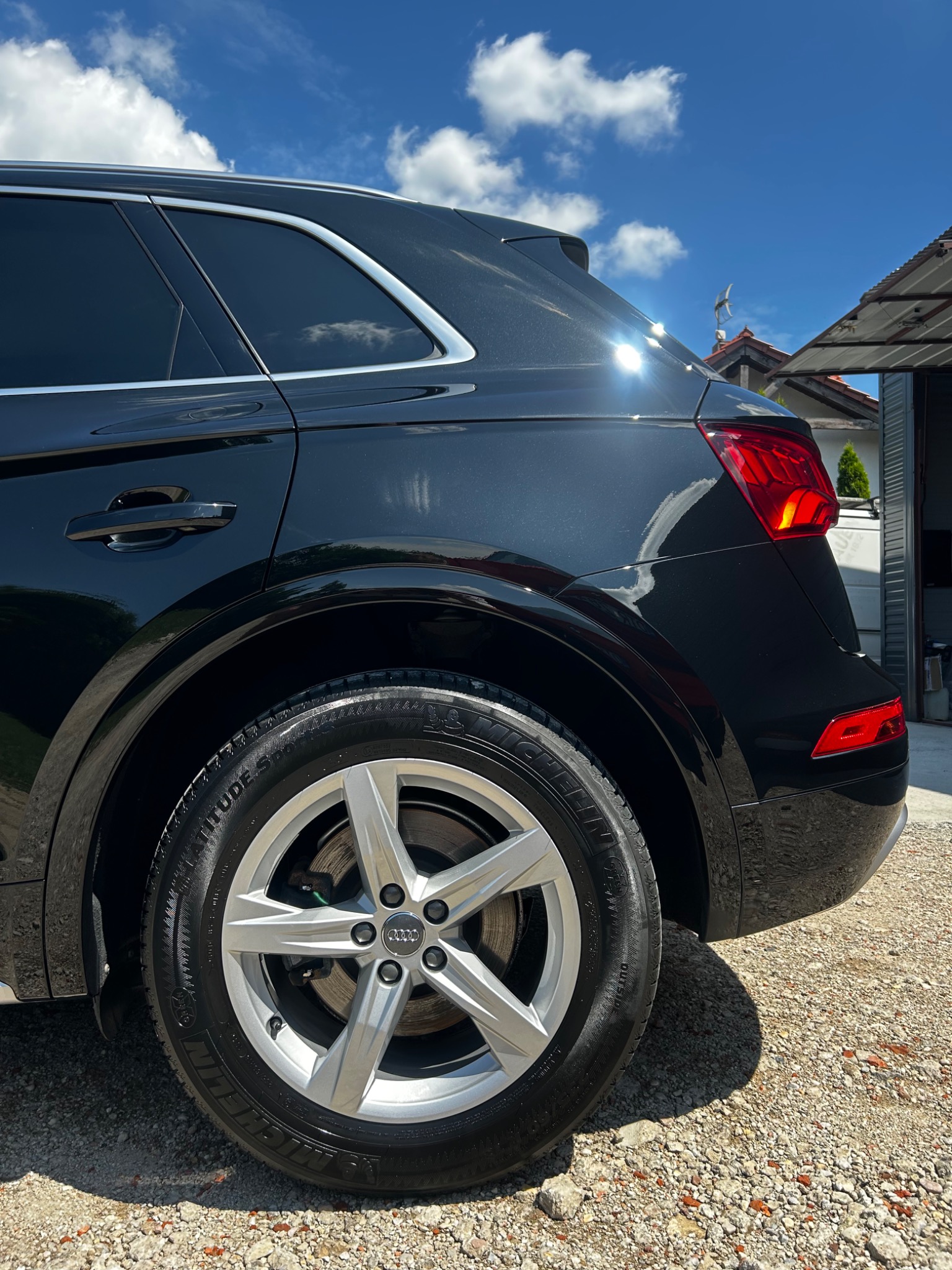 Błyszczący, czarny SUV Audi Q5 po umyciu, widoczny z tyłu pod kątem, z wyraźnie czystymi oponami Michelin i aluminiowymi felgami na tle błękitnego nieba z chmurami.