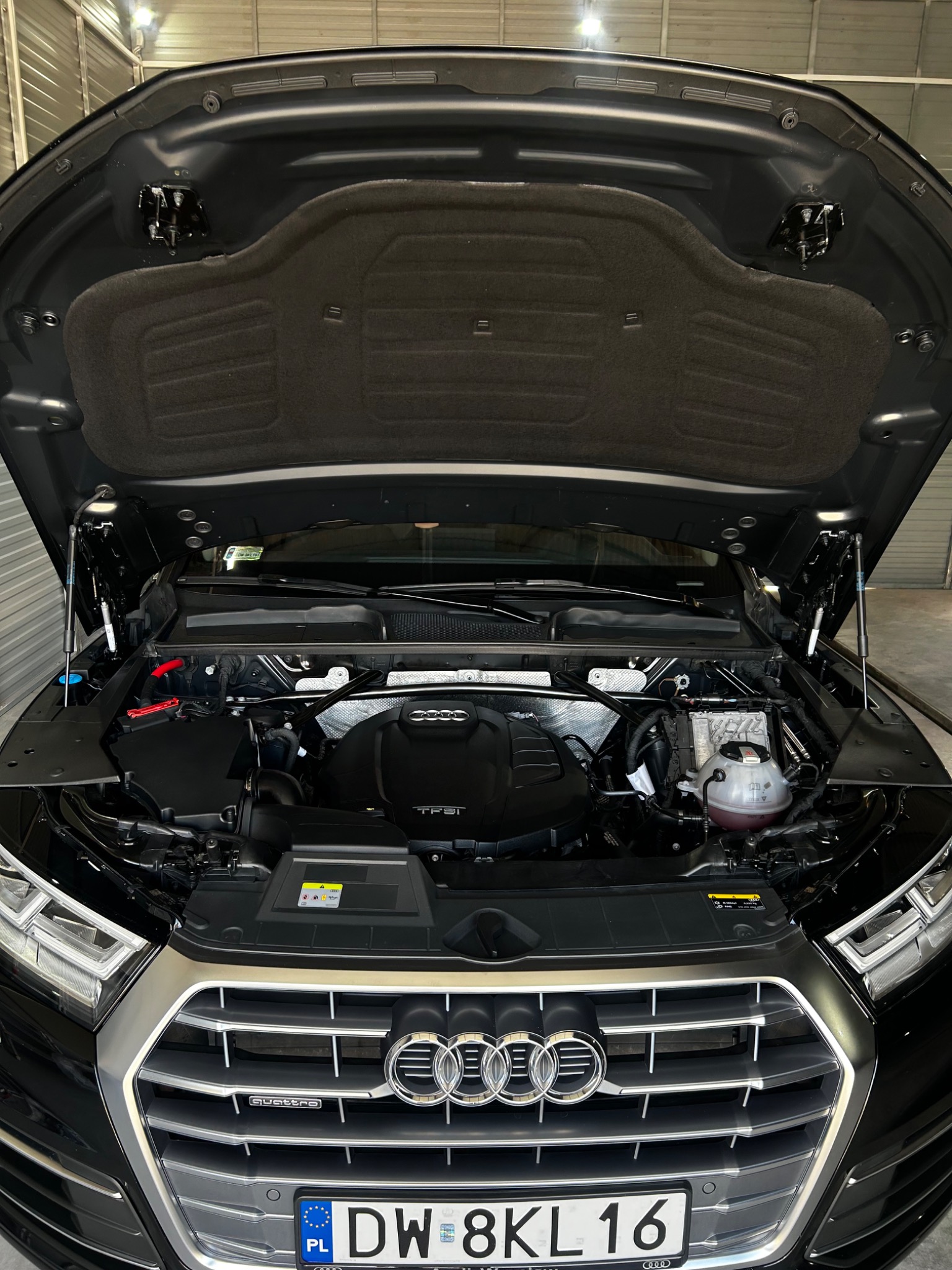 Otwarta maska czarnego samochodu Audi Q5 w Księginicach, widoczny silnik, elementy układu chłodzenia i tablica rejestracyjna.