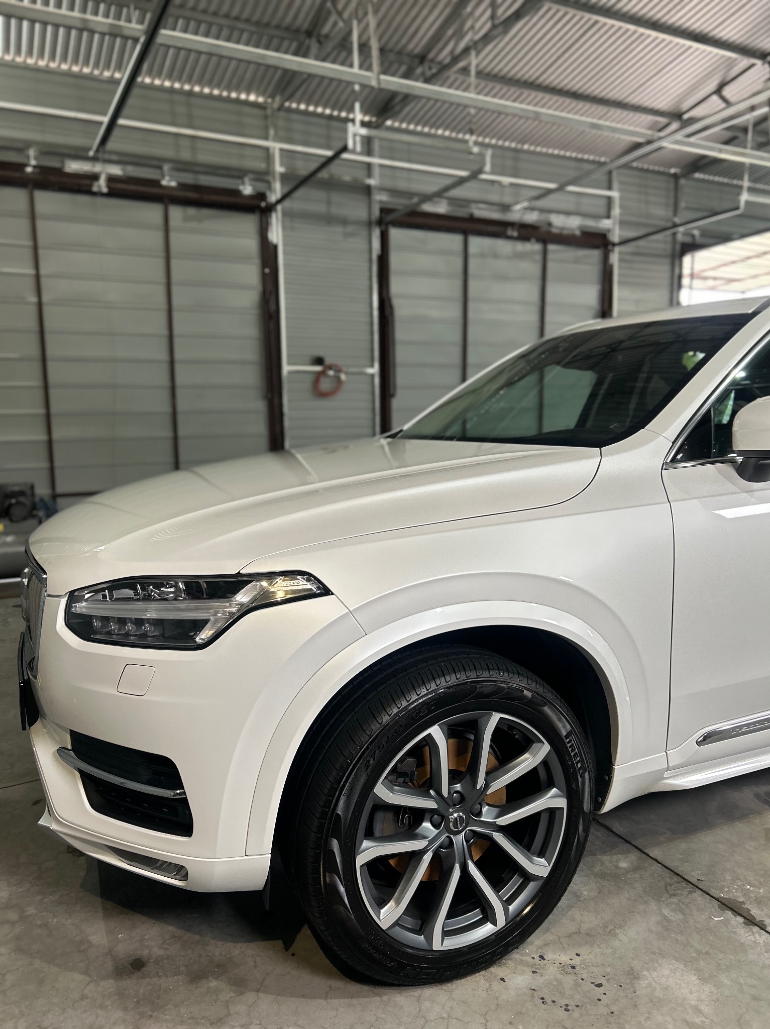 Biały samochód Volvo XC90 po umyciu, z błyszczącymi oponami i felgami, widoczny w jasnym garażu z metalowymi drzwiami.