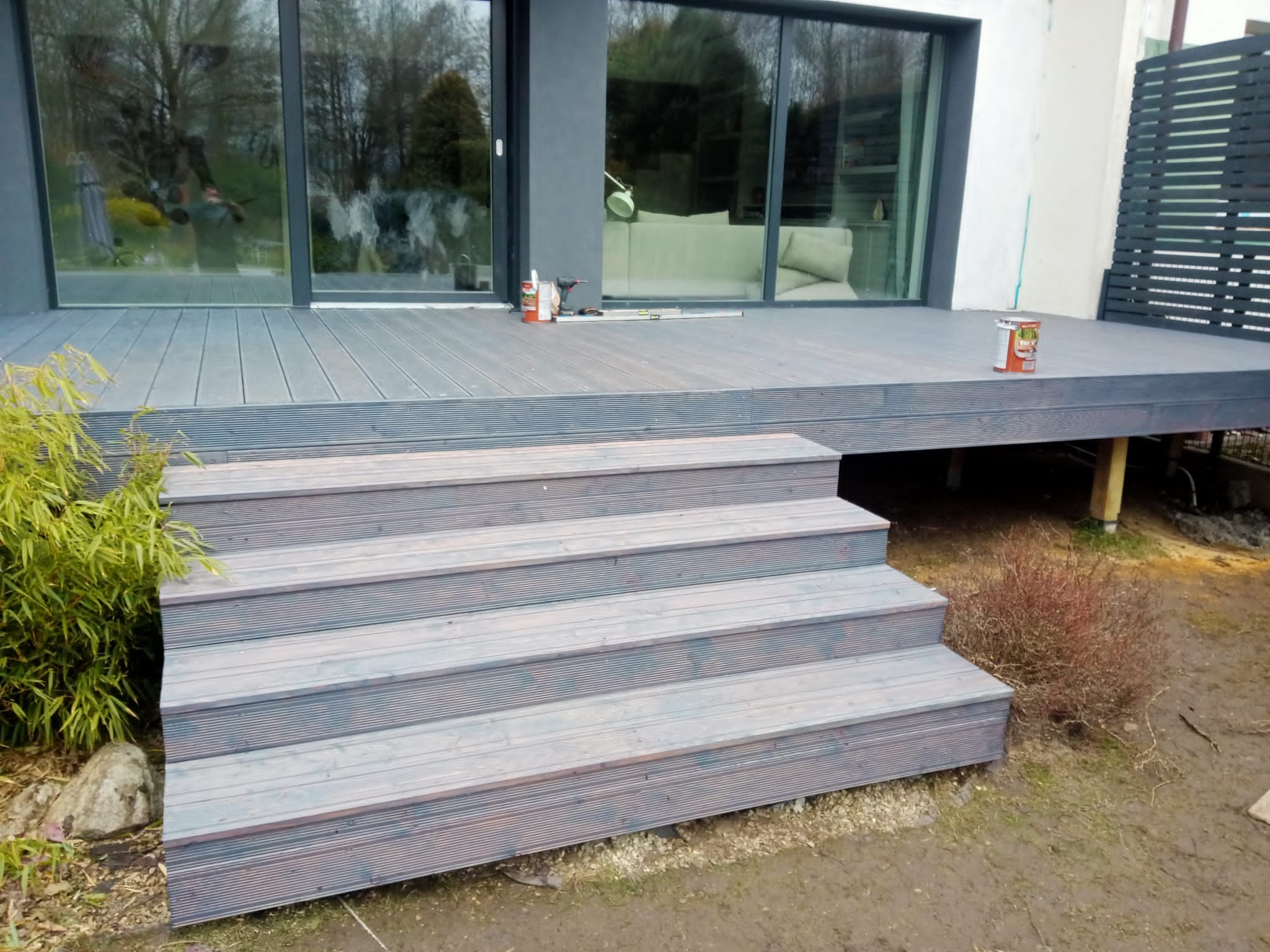 Nowoczesny taras z kompozytu w kolorze szarym, z niskimi schodami, widok na duże okno domu i narzędzia pracy.
