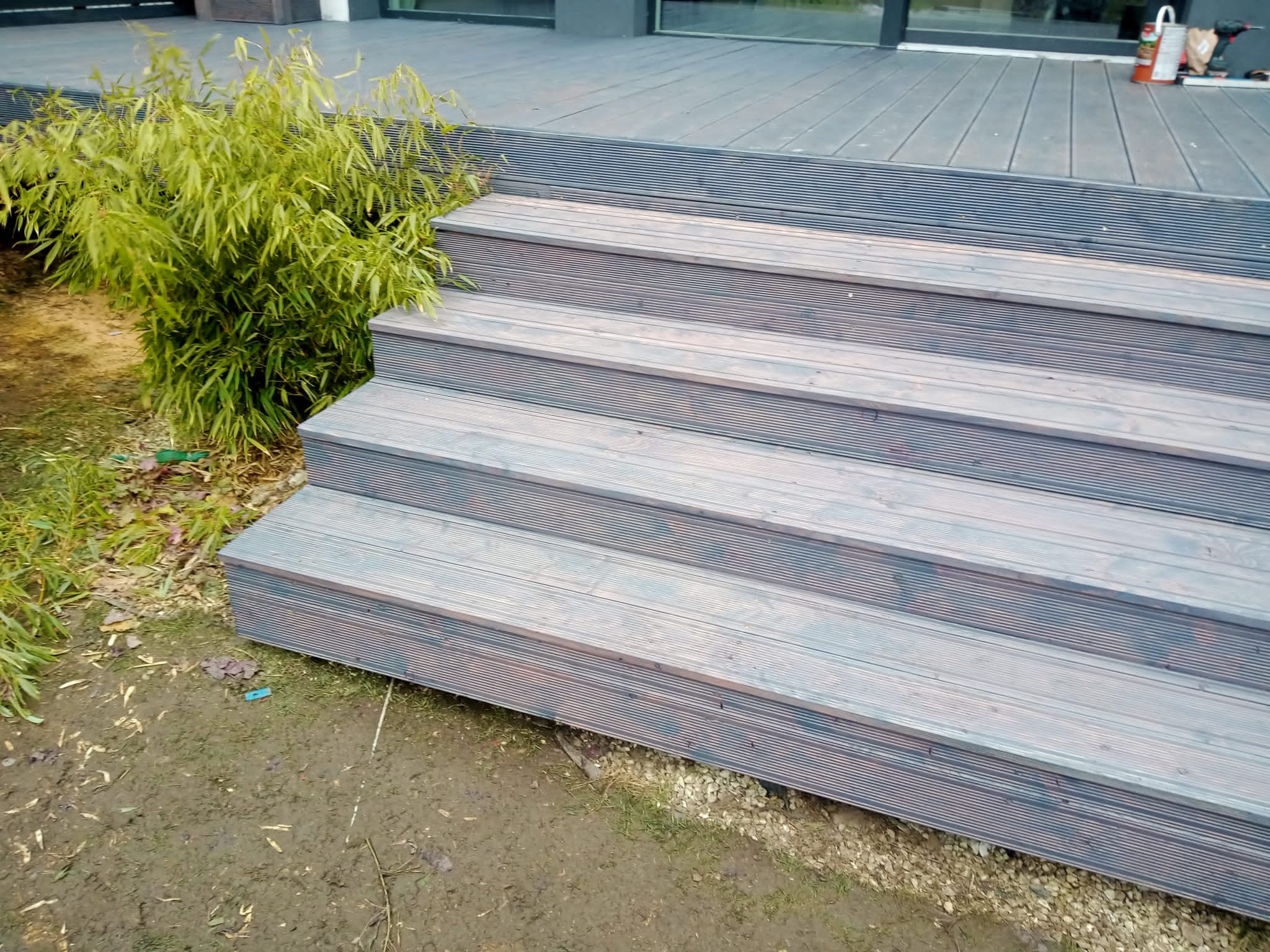 Schody z desek kompozytowych prowadzące na taras, obok rosnąca trawa ozdobna, widoczny fragment budynku.