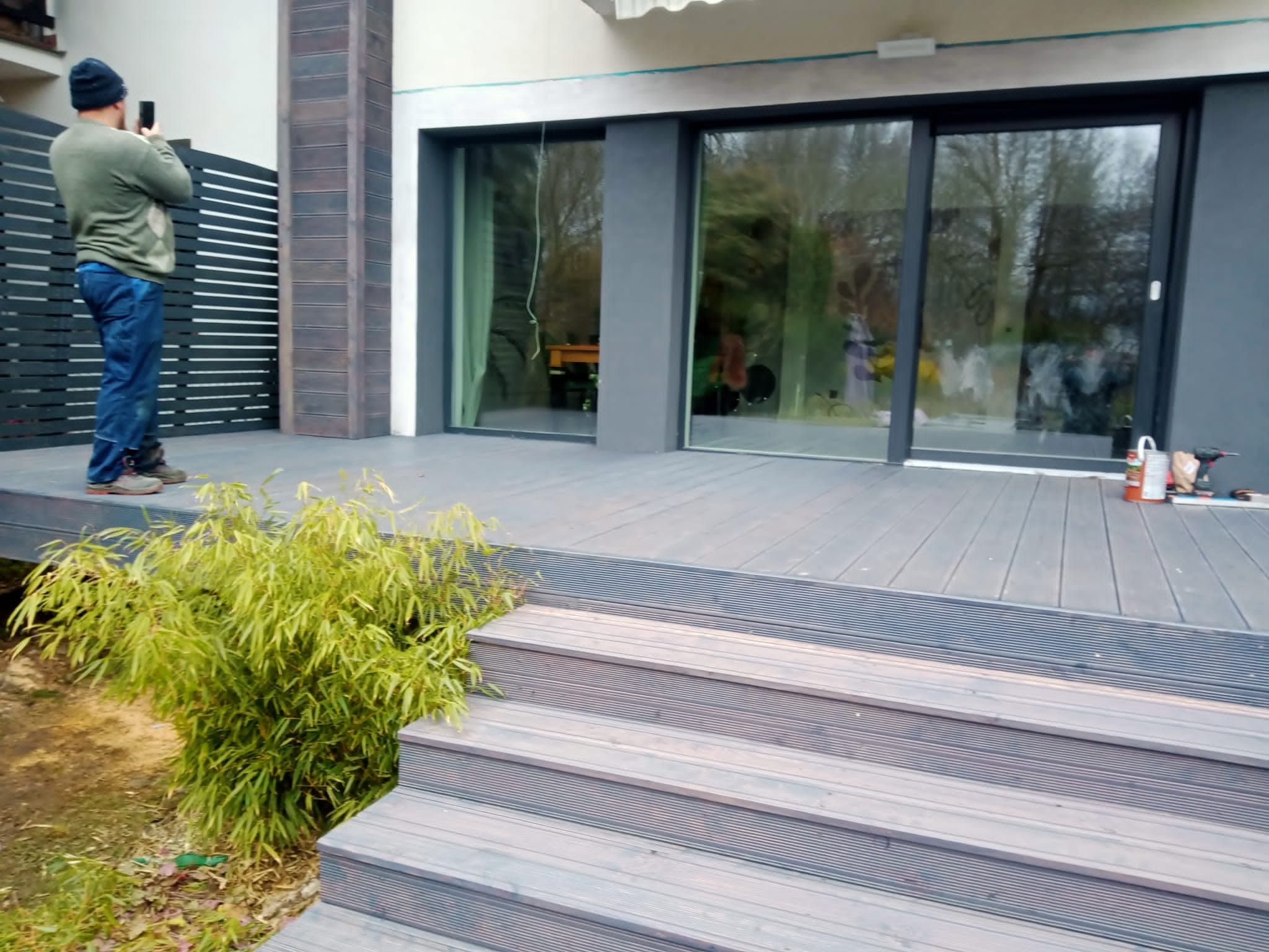 Mężczyzna fotografuje telefonem taras kompozytowy z niskimi schodami i ozdobną rośliną, widoczne narzędzia i przesuwne okna domu.