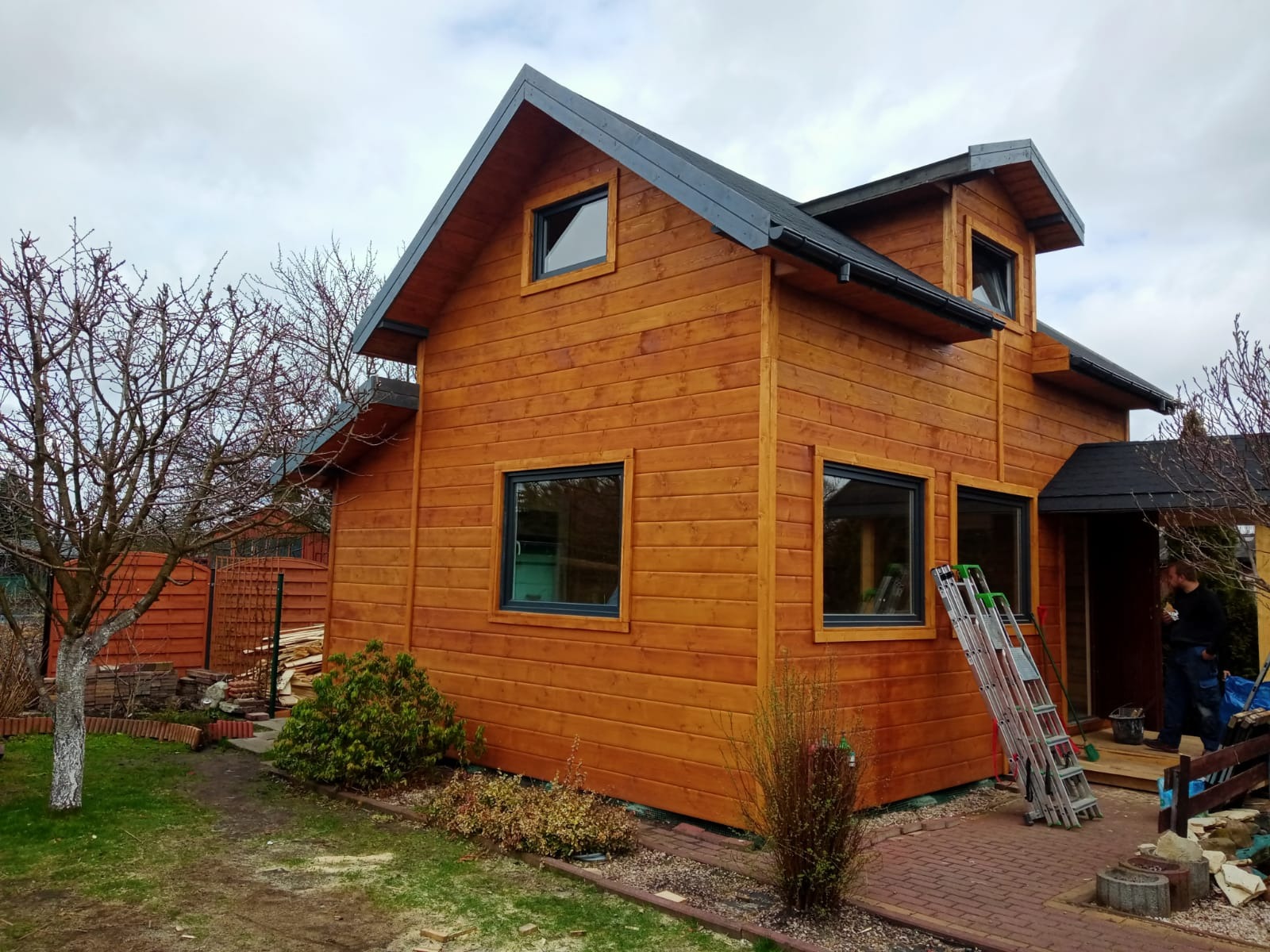 Zewnętrzny widok nowo wybudowanego, dwukondygnacyjnego domu z drewna, z ciemnymi oknami i dachem, obok którego stoi rozłożona drabina.