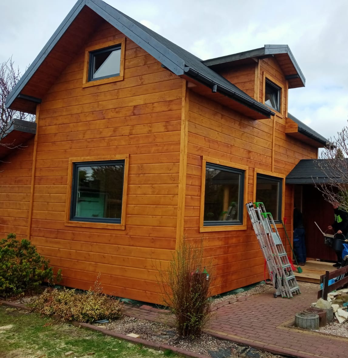 Nowoczesny, dwukondygnacyjny dom z drewna o ciepłym, brązowym odcieniu, z ciemnoszarą stolarką okienną i dachem, częściowo widoczna drabina oparta o elewację, kostka brukowa i zieleń wokół.