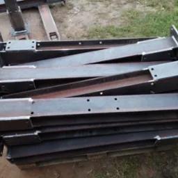 Stalowe elementy konstrukcyjne z otworami, ułożone warstwami na palecie, przygotowane do transportu lub montażu.