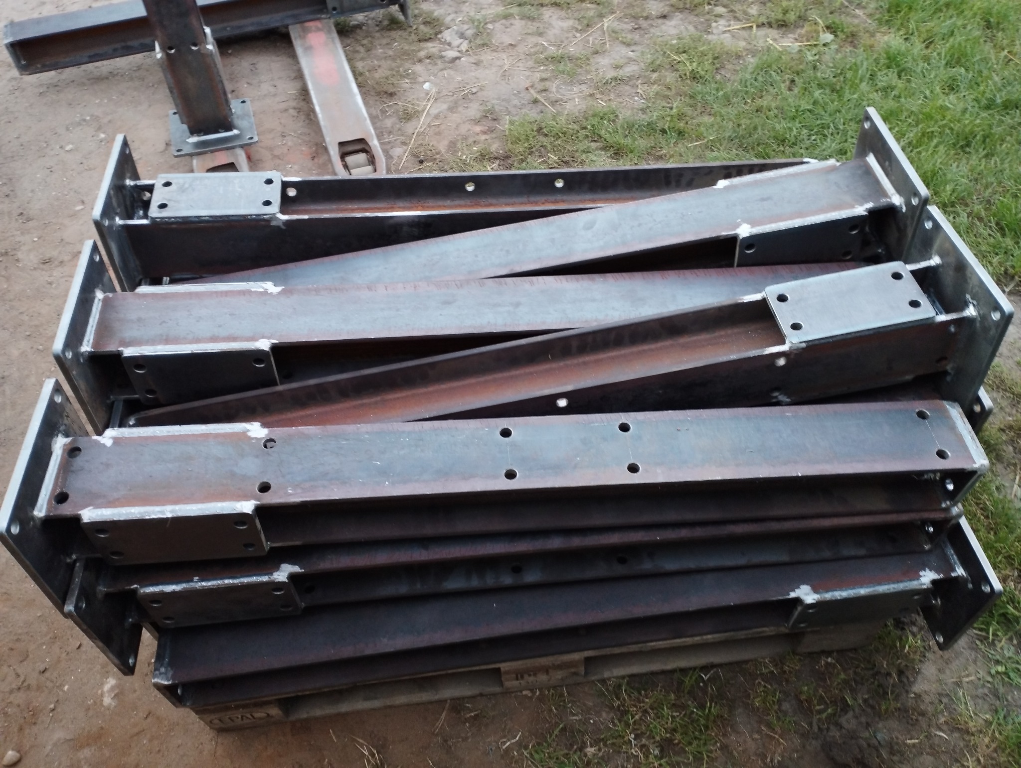 Stalowe elementy konstrukcyjne z otworami, ułożone warstwami na palecie, przygotowane do transportu lub montażu.