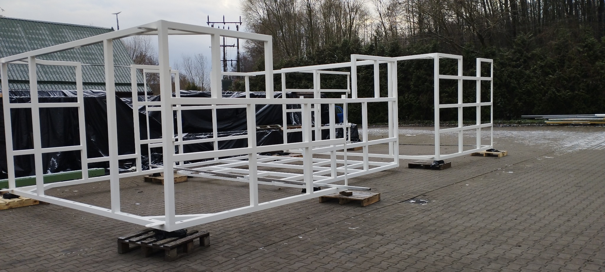 Biała, pomalowana proszkowo, modułowa konstrukcja stalowa na paletach, przygotowana do transportu, z elementami składowymi w tle i zielenią w tle.