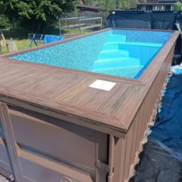 Nowoczesny, częściowo wkopany basen z mozaiką w odcieniach błękitu i schodami, otoczony drewnianym tarasem w kolorze brązowym, widok z góry na tle zieleni.