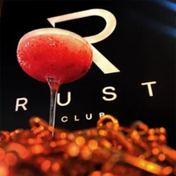 Kieliszek z czerwonym koktajlem ozdobionym drobnymi kuleczkami na tle logo 'RUST CLUB' i dekoracyjnych, rozmytych elementów w odcieniach pomarańczy.