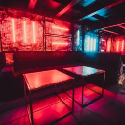 Wnętrze klubu nocnego z neonowym oświetleniem w odcieniach czerwieni i niebieskiego, ciemnoszara sofa i stoliki z metalowymi ramami.