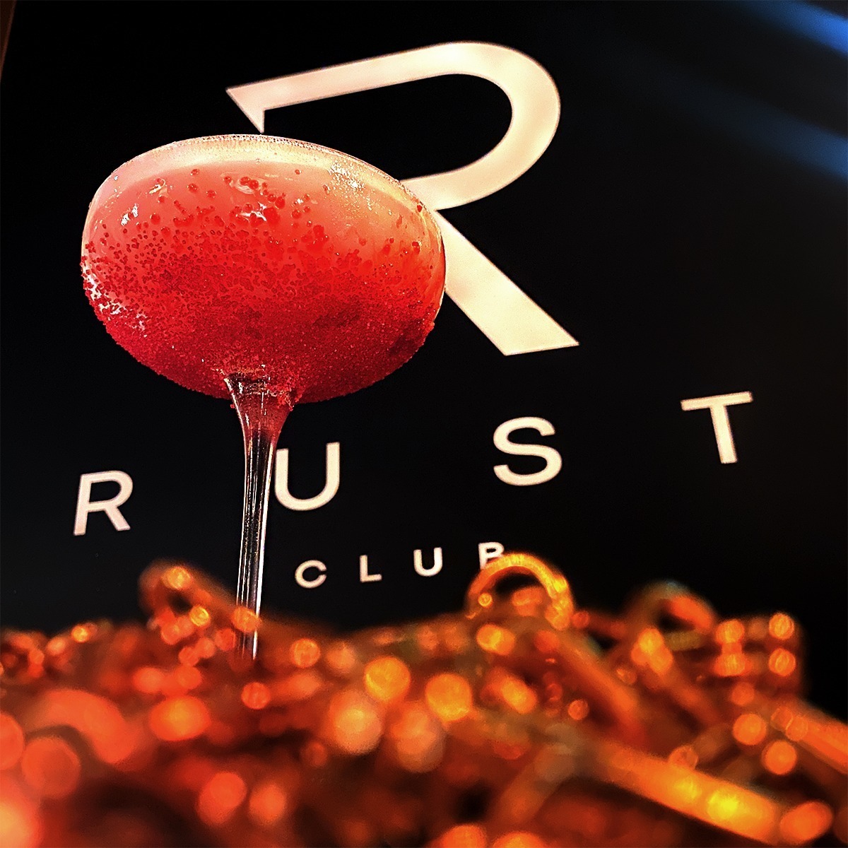 Kieliszek z czerwonym koktajlem ozdobionym drobnymi kuleczkami na tle logo 'RUST CLUB' i dekoracyjnych, rozmytych elementów w odcieniach pomarańczy.