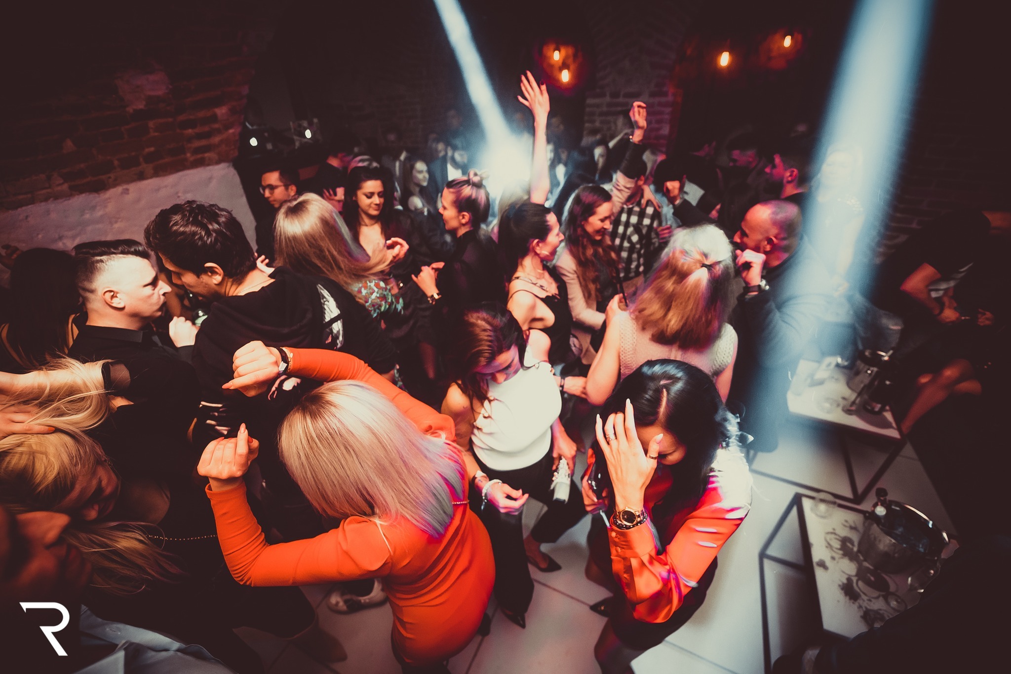 Tłum ludzi tańczących w klubie nocnym z ceglanymi ścianami i oświetleniem punktowym, widoczne stoliki z napojami.