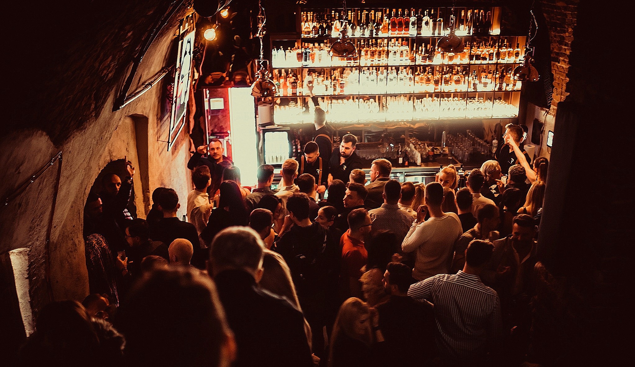Tłum ludzi w klimatycznym barze z ceglanym sklepieniem, oświetlonym rzędami butelek z alkoholem i wiszącymi lampami, barmani za barem przygotowują drinki.
