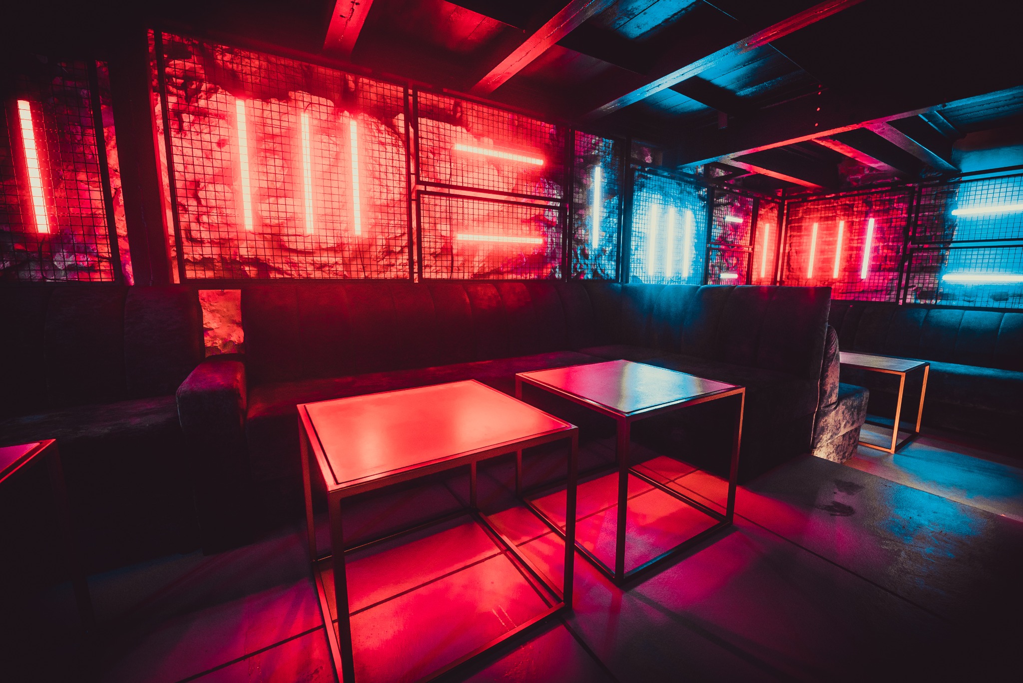 Wnętrze klubu nocnego z neonowym oświetleniem w odcieniach czerwieni i niebieskiego, ciemnoszara sofa i stoliki z metalowymi ramami.
