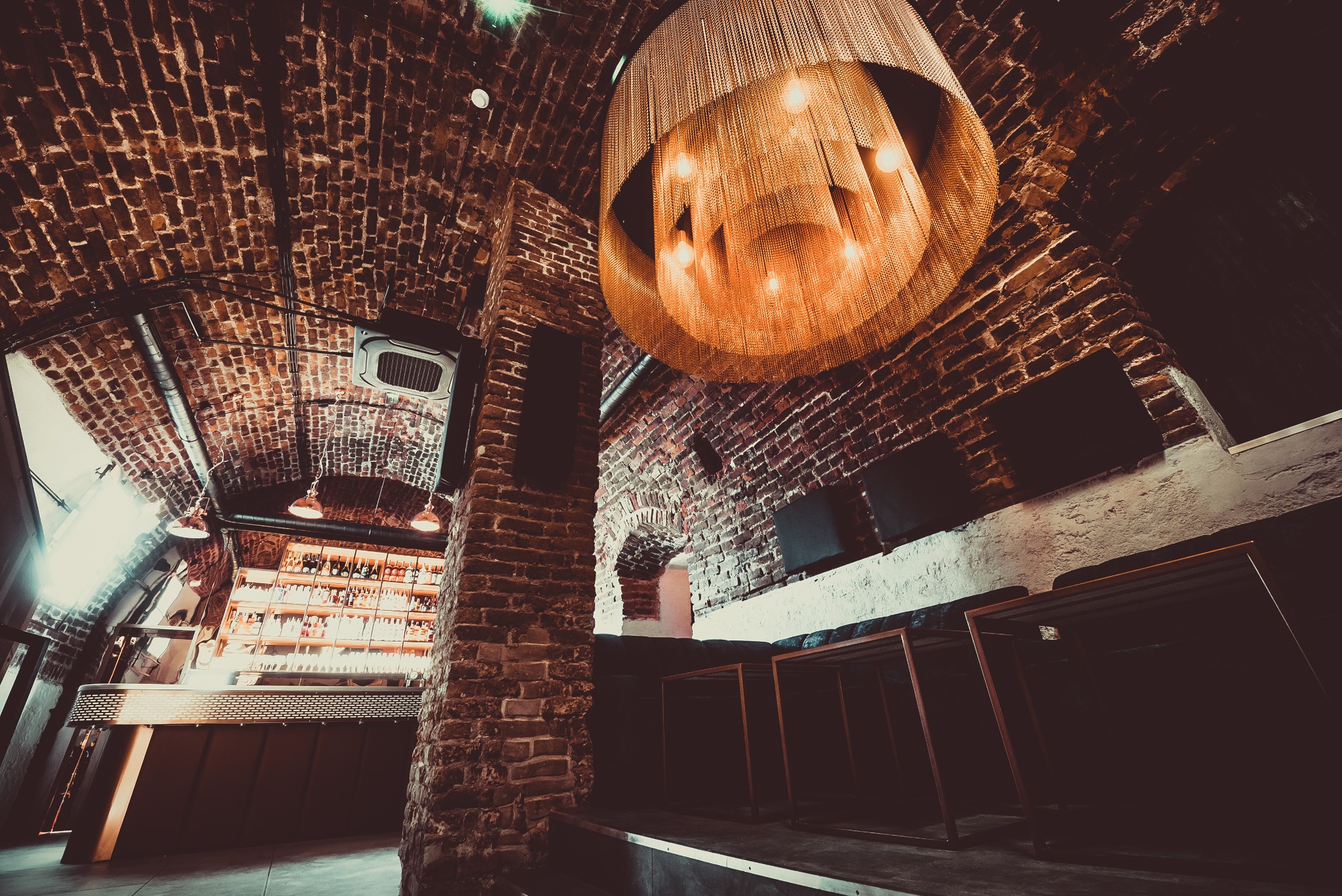 Wnętrze lokalu z ceglanym sklepieniem, ozdobnym żyrandolem z łańcuszków, barem z podświetlonymi półkami pełnymi butelek oraz lożą z niskimi stolikami.