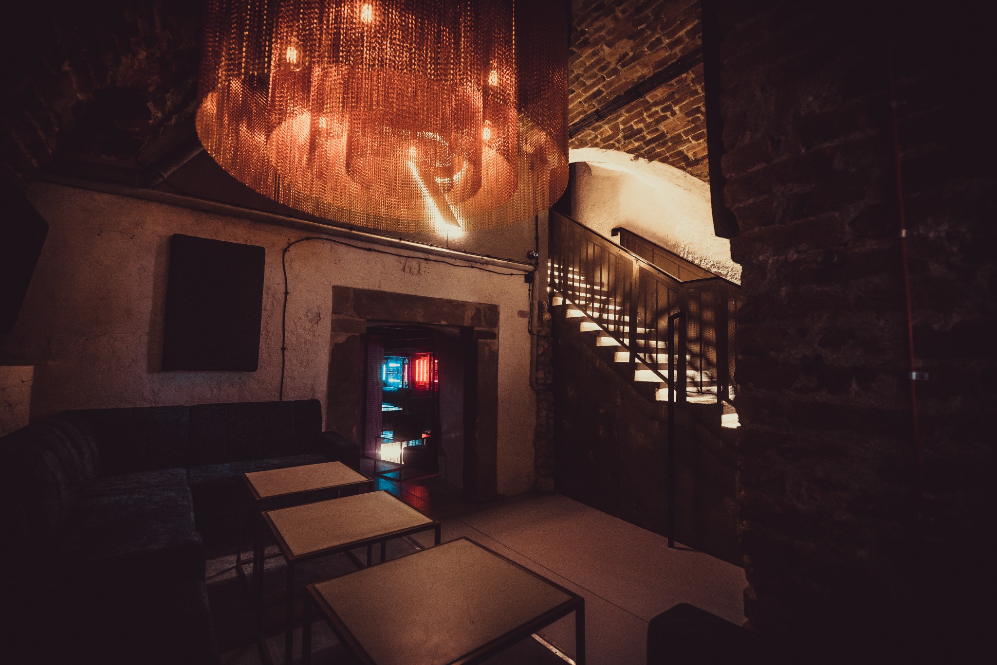 Wnętrze klubu z ceglanymi ścianami, metalową lampą w kształcie kuli, schodami z podświetleniem i sofą z niskimi stolikami.