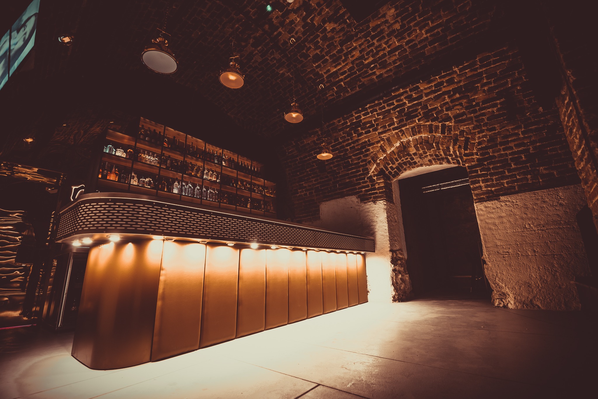 Oświetlony bar z podświetlanym frontem, półkami pełnymi butelek i ceglanym łukiem w tle, w ciemnym wnętrzu.