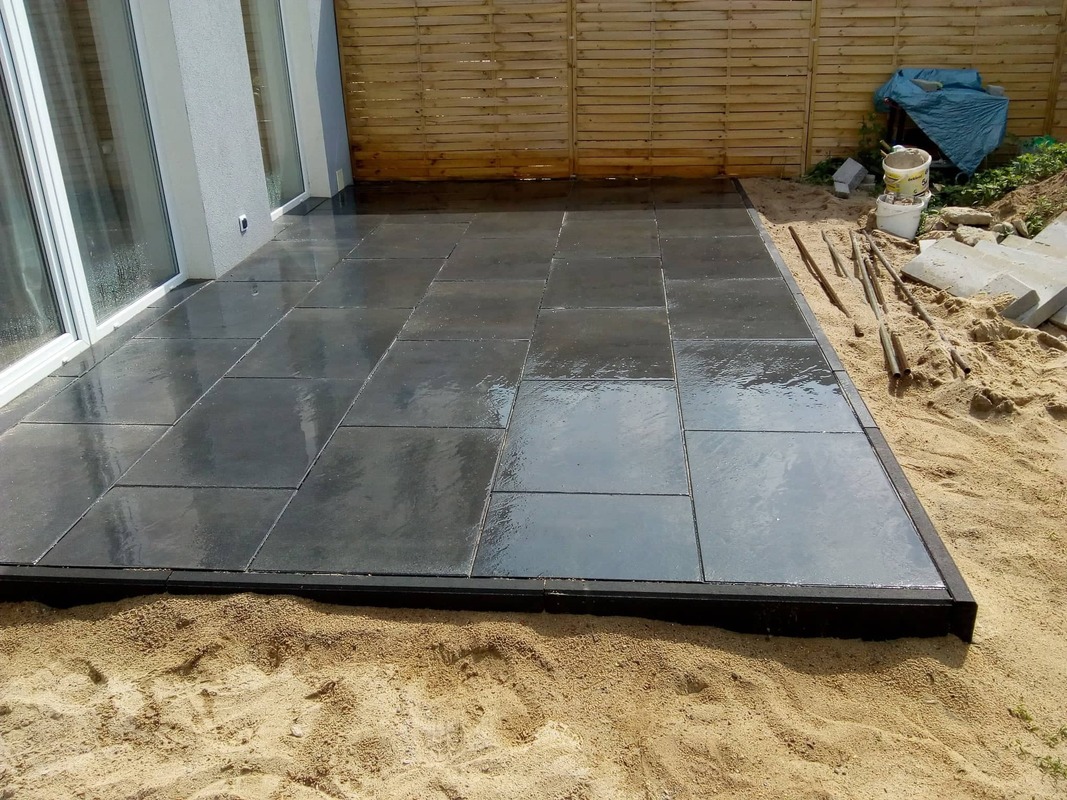Świeżo ułożony, lśniący taras z ciemnoszarej kostki brukowej na piaskowym podłożu, z widocznym obrzeżem i elementami przygotowania do dalszych prac w tle.