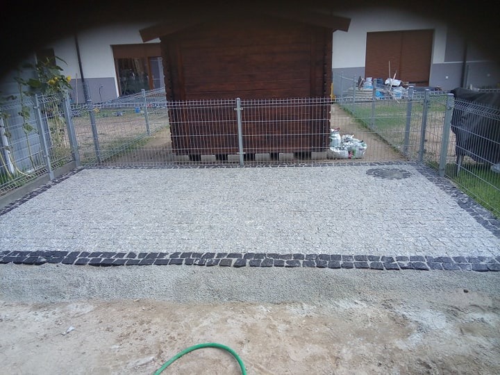 Niewielki plac wyłożony jasną kostką z obramowaniem z ciemnej kostki granitowej, otoczony metalowym ogrodzeniem i drewnianą szopą w tle, widok z góry.