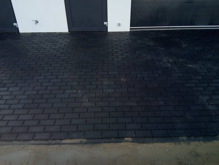 Świeżo ułożona, czarna kostka brukowa przed garażem z białymi ścianami i czarnymi drzwiami, widoczne ślady wilgoci na powierzchni.