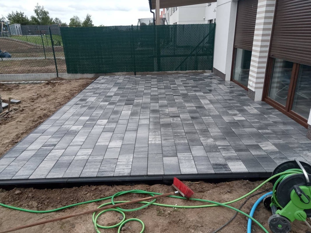 Ułożona szara kostka brukowa na posesji przy domu jednorodzinnym, widoczne narzędzia i zwinięty wąż ogrodowy.