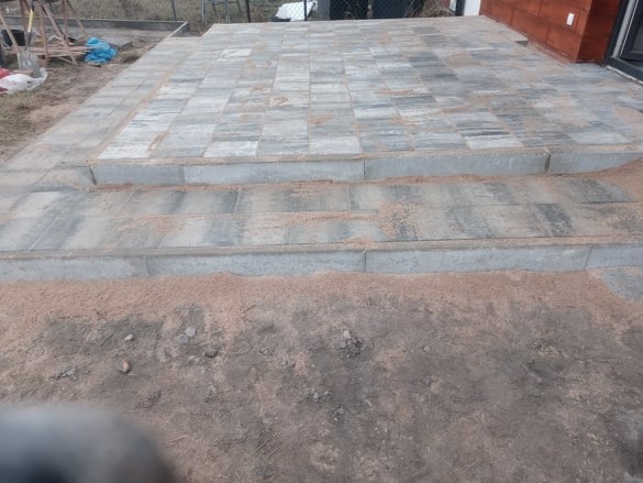 Ułożona szara kostka brukowa tworząca taras z dwustopniowymi schodami, zasypana piaskiem kwarcowym, widok z poziomu gruntu.