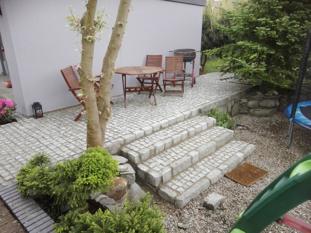 Granitowe schody prowadzące na taras z kostki granitowej, na którym znajdują się drewniane meble ogrodowe i grill.