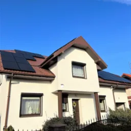 Biały dom jednorodzinny z brązowym dachem i panelami słonecznymi na dachu, widok z dołu na tle błękitnego nieba.