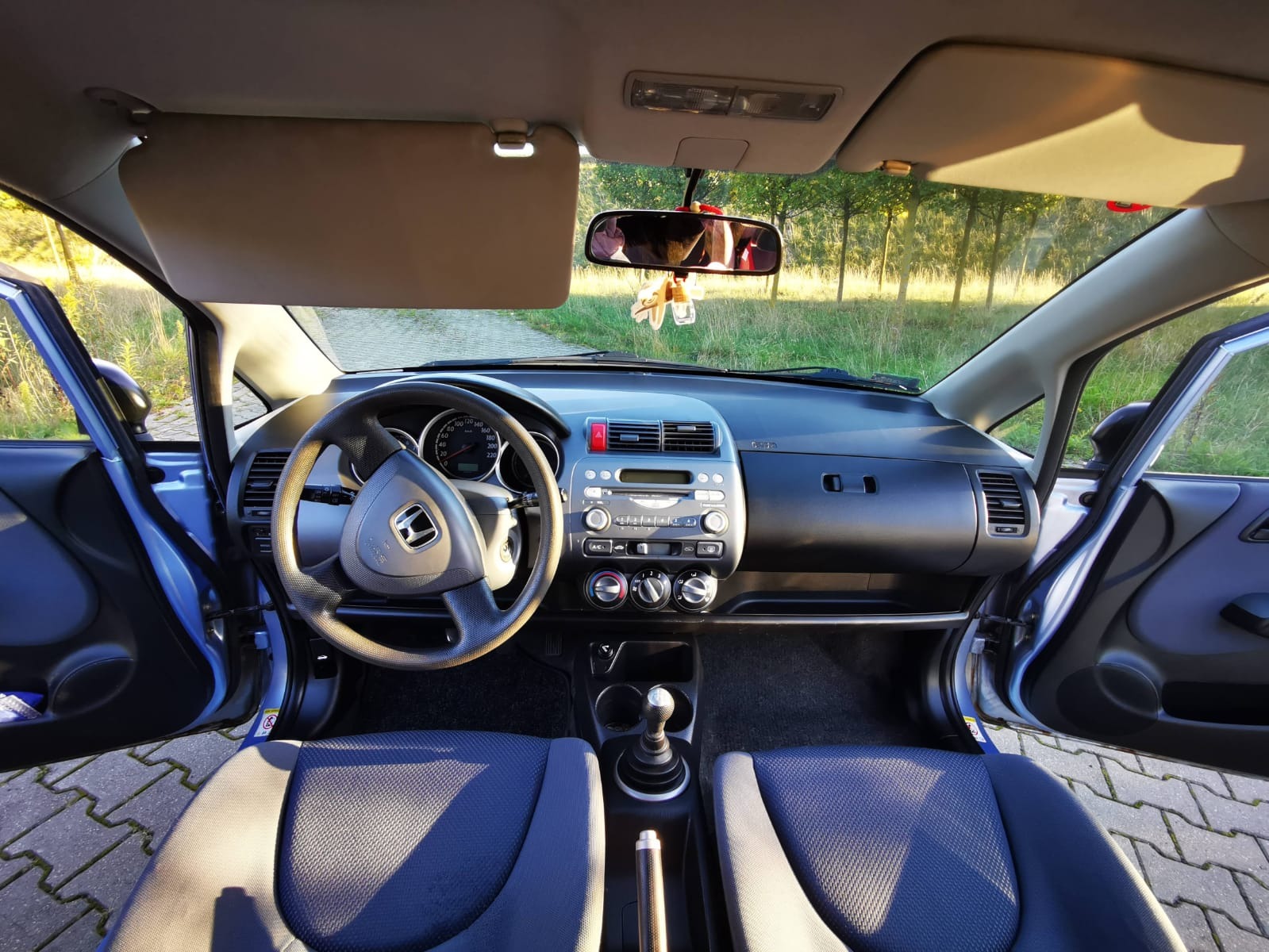 Wnętrze samochodu Honda Jazz z otwartymi drzwiami, widok na deskę rozdzielczą, kierownicę z logo, niebieskie fotele i fragment drogi na zewnątrz.