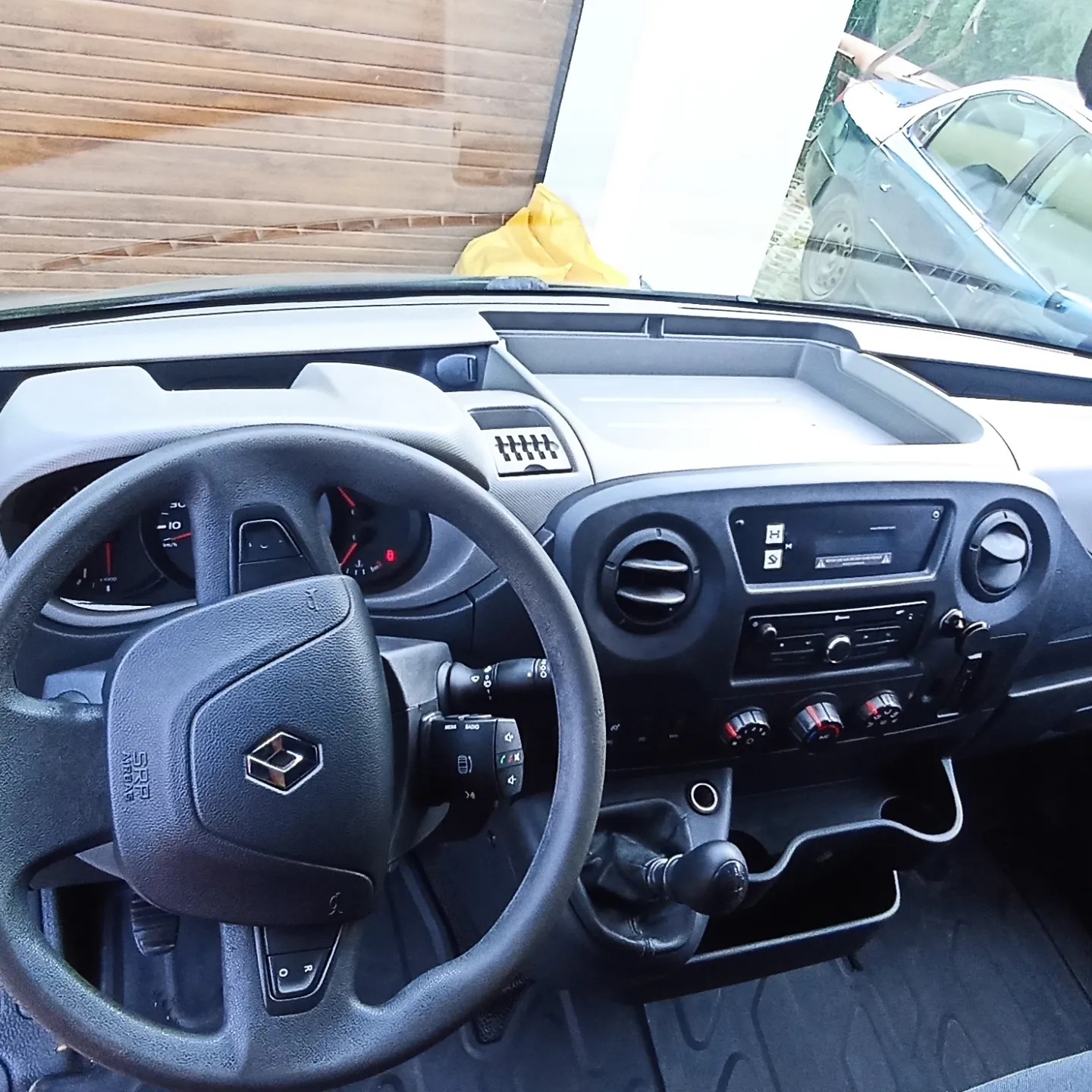 Wnętrze samochodu dostawczego Renault Master, widok na deskę rozdzielczą, kierownicę z logo, radio i dźwignię zmiany biegów. Na zewnątrz widoczny fragment innego pojazdu.