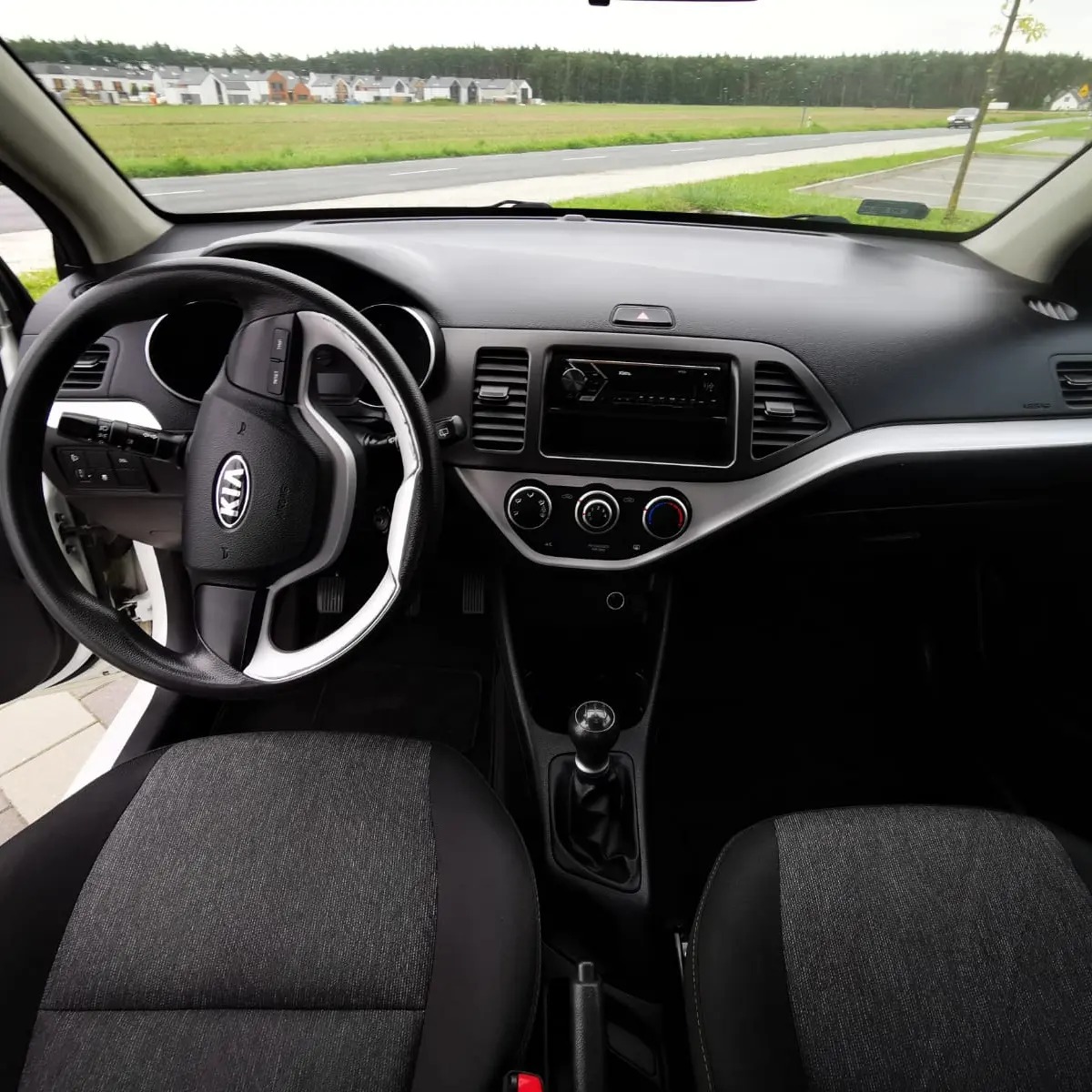Wnętrze samochodu Kia Picanto z widocznymi fotelami, deską rozdzielczą i kierownicą z logo marki, w tle widok na drogę i zabudowania.