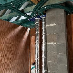 Wentplast - Zielone, karbowane rury wentylacyjne przymocowane do sufitu i podłączone do srebrnych, izolowanych rur pionowych w surowym wnętrzu budynku z ceglanymi ścianami.