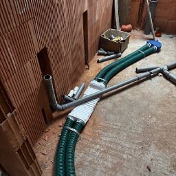 Wentplast - Montaż rekuperacji w surowym wnętrzu: zielone rury wentylacyjne podłączone do wymiennika ciepła, widoczne elementy instalacji i ściany z cegły dziurawki.