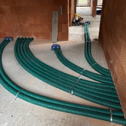 Wentplast - Zielone rury wentylacyjne ułożone na podłodze w surowym wnętrzu budynku z czerwonej cegły, przygotowane do montażu systemu rekuperacji.