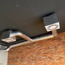 Wentplast - Dwa srebrne przewody wentylacyjne z białymi kratkami na suficie, na tle ceglanej ściany. Widoczny klimatyzator. Ujęcie z dołu, podkreślające instalację.