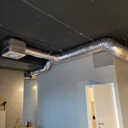 Wentplast - Srebrne, izolowane rury wentylacyjne zamontowane pod ciemnym sufitem w surowym wnętrzu. Widoczny anemostat i fragment ściany z szarych płytek.
