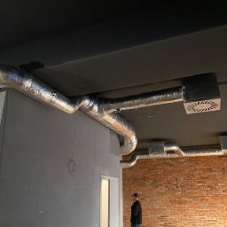 Wentplast - Montaż instalacji wentylacyjnej z aluminiowymi rurami na suficie w surowym wnętrzu. Widoczny fragment ceglanej ściany i osoba w oddali.