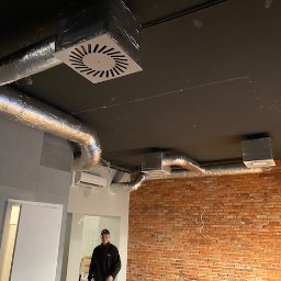 Wentplast - Zamontowana instalacja wentylacyjna z aluminiowymi rurami i kratkami na suficie w pomieszczeniu z ceglaną ścianą. Widoczny klimatyzator i osoba w tle.