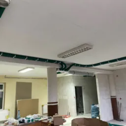 Zielone, karbowane rury wentylacyjne z białymi uchwytami zamontowane pod sufitem w pomieszczeniu w trakcie remontu, widoczne lampy sufitowe i fragmenty ścian z cegieł. Na pierwszym planie widoczny...
