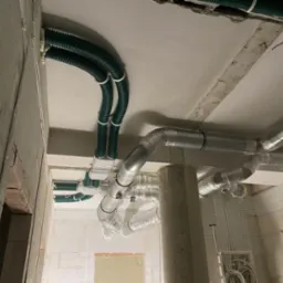 Wnętrze w trakcie instalacji rekuperacji i wentylacji: widoczne zielone karbowane rury wentylacyjne podwieszone pod sufitem, aluminiowe rury spiro, oraz liczne kable elektryczne prowadzone po...