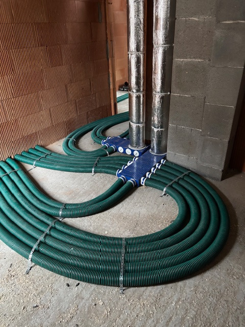 Rozprowadzona instalacja rekuperacji z zielonymi rurami i niebieskimi rozdzielaczami, widoczne dwa pionowe, zaizolowane przewody. Surowy stan budynku.