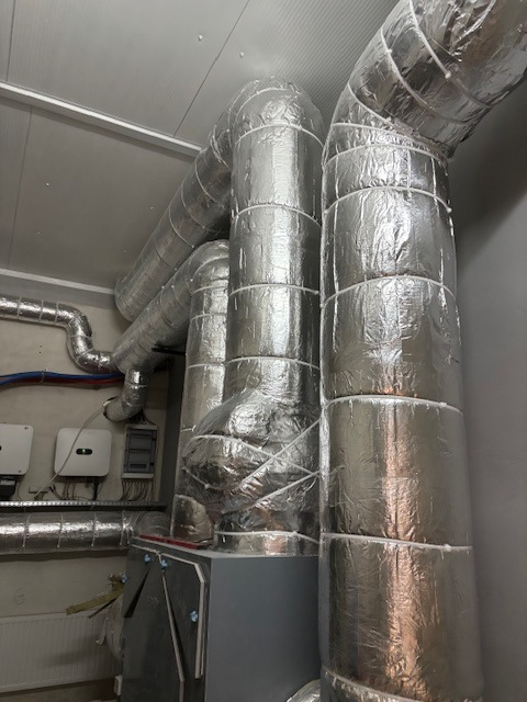 Izolowane rury wentylacyjne w pomieszczeniu technicznym, widoczne panele sterowania i inne instalacje. Srebrna izolacja na rurach, szare ściany i sufit.