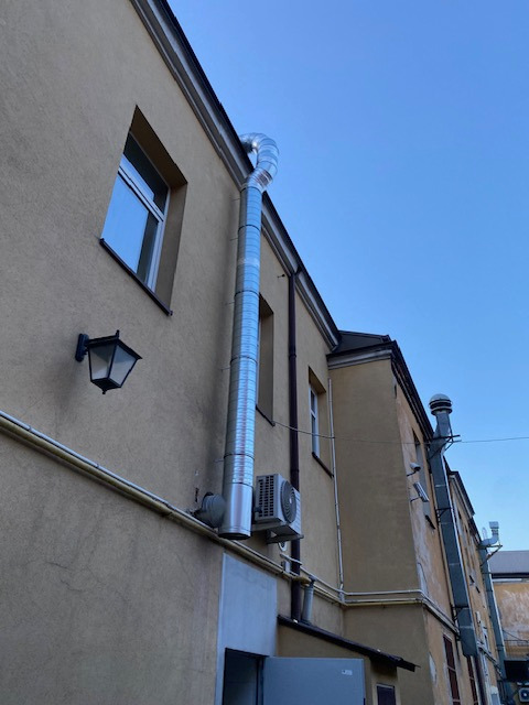 Zewnętrzna instalacja wentylacyjna z rurami spiro na elewacji budynku w Kielcach. Widoczny fragment okna i lampy. Ujęcie z dołu, błękitne niebo w tle.