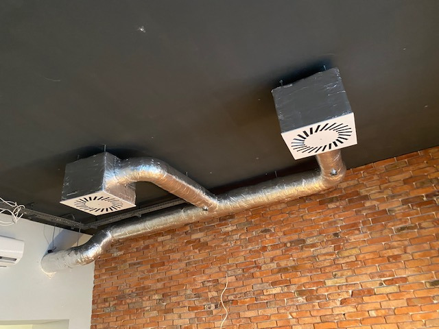 Dwa srebrne przewody wentylacyjne z białymi kratkami na suficie, na tle ceglanej ściany. Widoczny klimatyzator. Ujęcie z dołu, podkreślające instalację.