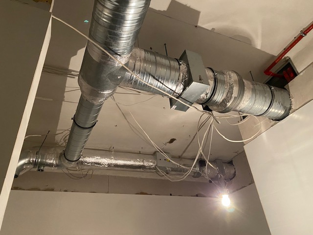 Srebrne, metalowe rury wentylacyjne pod sufitem, połączone kształtkami i izolacją. Widoczne przewody elektryczne i fragment instalacji przeciwpożarowej.
