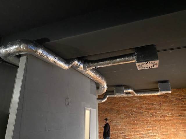 Montaż instalacji wentylacyjnej z aluminiowymi rurami na suficie w surowym wnętrzu. Widoczny fragment ceglanej ściany i osoba w oddali.