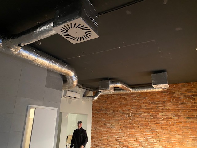 Zamontowana instalacja wentylacyjna z aluminiowymi rurami i kratkami na suficie w pomieszczeniu z ceglaną ścianą. Widoczny klimatyzator i osoba w tle.