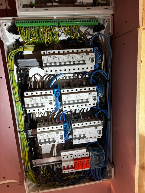 Rozdzielnica elektryczna w białej obudowie z widocznymi bezpiecznikami, przewodami w różnych kolorach i listwami zaciskowymi. Instalacja w nowym budynku.