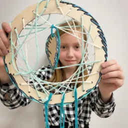 Dziewczynka prezentuje własnoręcznie wykonany łapacz snów z drewnianej obręczy, białego i turkusowego sznurka oraz piórka, z czarnym wzorem na obrzeżach i piórku.