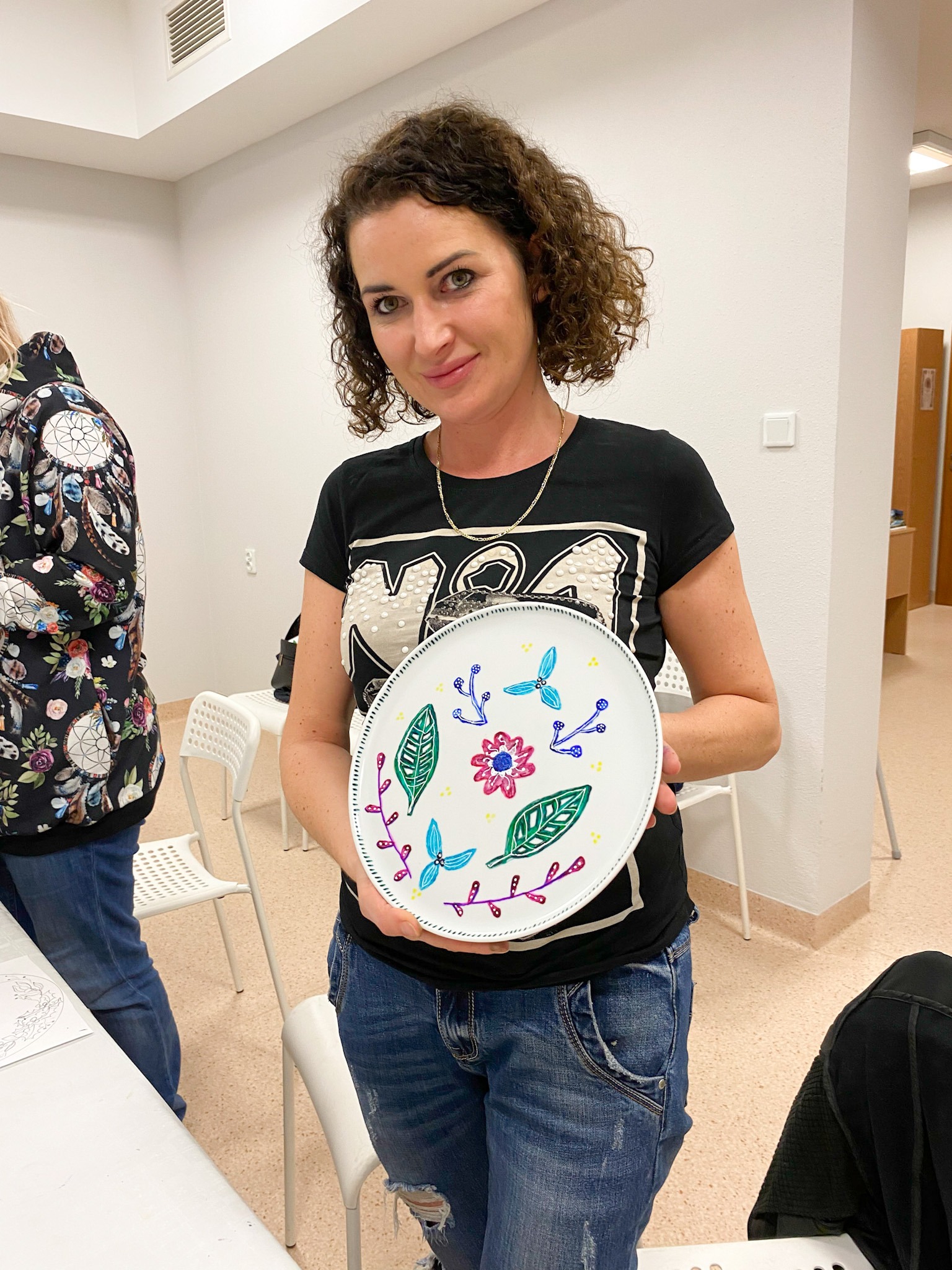 Uśmiechnięta kobieta z kręconymi włosami prezentuje biały talerz z ręcznie malowanymi kolorowymi motywami roślinnymi i kwiatowymi.